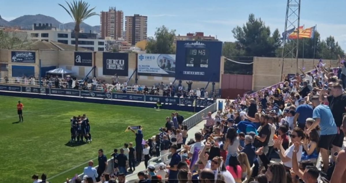 A pristine modern football stadium bowl seen from a high corner angle, with vivid green grass, bright blue seating and UK-style supporters’ flags draped over empty seats.